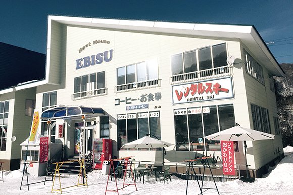 外壁に「Rest House EBISU」の看板がある白い建物。屋根が青。前にスキーラックとテラス席