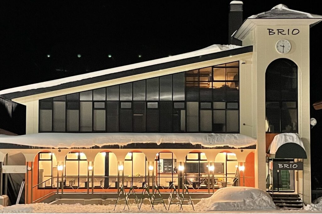 Rest House BRIO exterior: building with a clock tower, ski racks and skiers in front